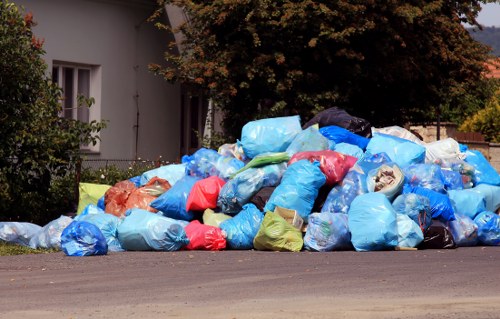 A large pile of rubbish bags, predominantly blue with some red, yellow, and black, is accumulated on the side of a paved road in a residential area. The bags appear to be made of plastic and are filled with household waste or similar rubbish, with some bags slightly bulging and others partially torn or open, revealing contents that include paper and light materials. Behind the pile, there is a white residential building with visible windows and a garden area with green foliage and a tree with dark green and brown leaves. The outdoor setting is illuminated by natural daylight, and the rubbish pile is situated close to the footpath, indicating a rubbish removal or clearance activity, possibly performed by a local waste management service such as Office Clearance Havering, in the Havering area. The scene emphasizes the presence of accumulated waste awaiting removal, set against the backdrop of suburban Havering's residential environment.