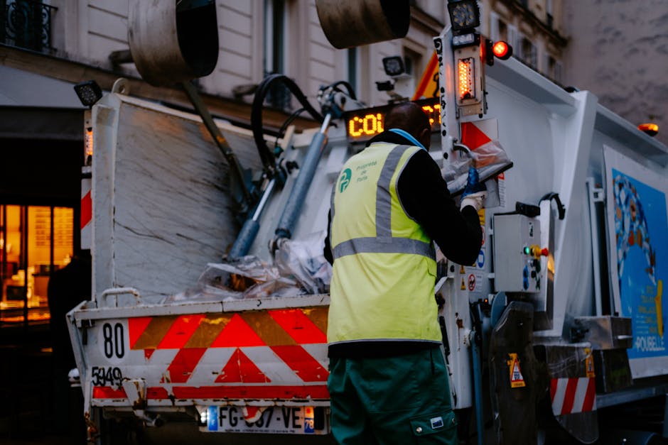 A large collection of mixed rubbish including flattened cardboard boxes, plastic bottles, paper bags, and discarded packaging is piled around multiple waste bins and containers on a paved urban street