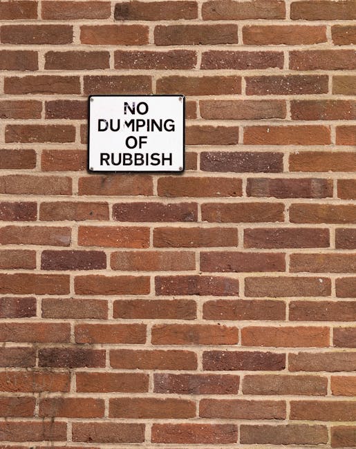 A rectangular white metal sign with black uppercase text reading 'NO DUMPING OF RUBBISH' is mounted on a red-brown brick wall. The bricks are arranged in a typical running bond pattern, with some dark