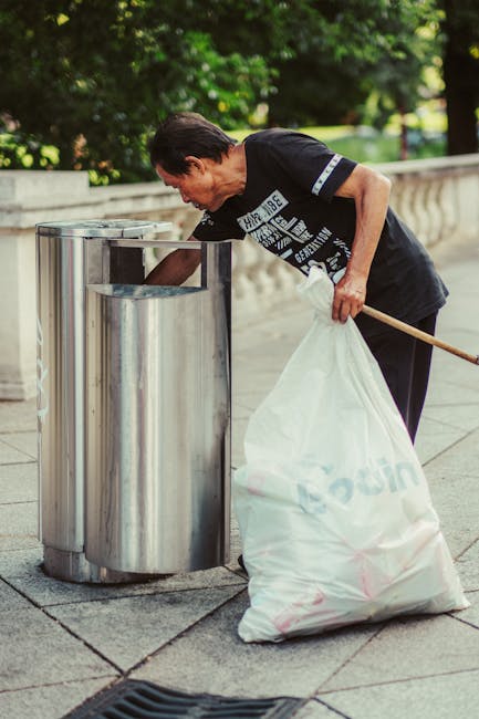 A waste collection worker wearing a green uniform and safety helmet is operating a large, industrial rubbish truck on a quiet street lined with trees and parked cars. The truck has a rear loading mech