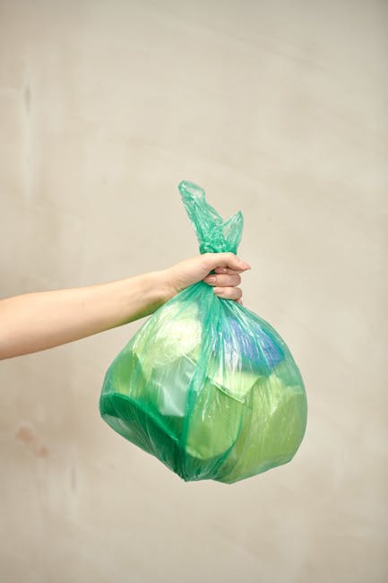 A person's hand and forearm are visible, holding a green plastic rubbish bag tightly at the top, with the knot gathered into a small twist tie. The bag appears full, slightly bulging, and contains mix