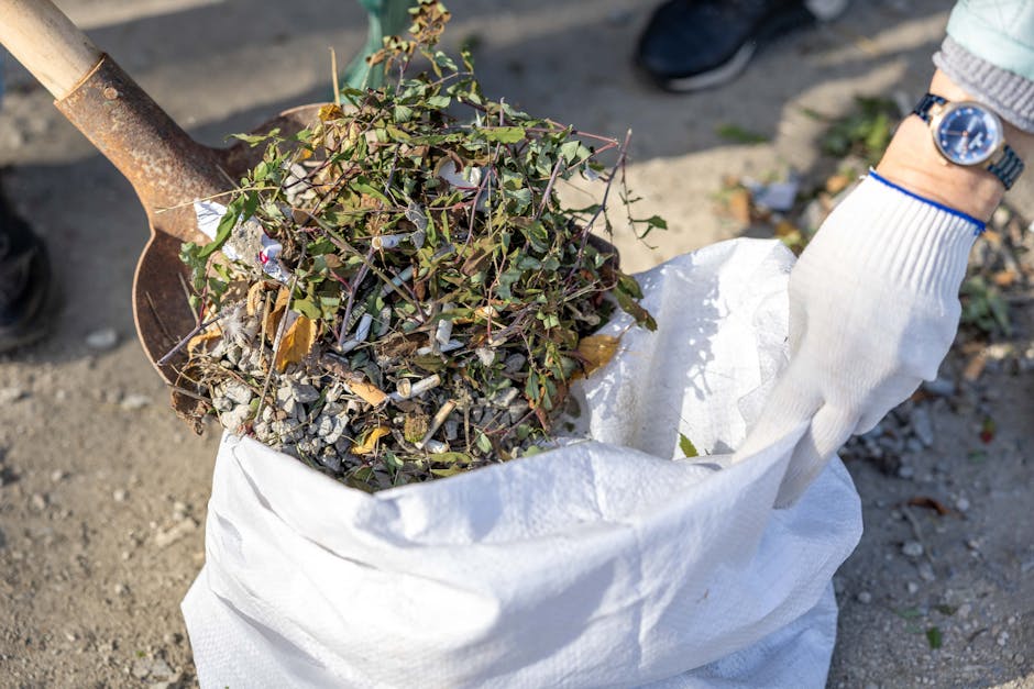A person wearing a white glove is holding open the top of a large white heavy-duty sack filled with garden waste, including a mix of small branches, leaves, and plant debris. The garden waste consists