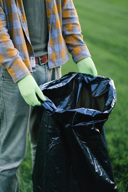 A person wearing a white glove is holding open the top of a large white heavy-duty sack filled with garden waste, including a mix of small branches, leaves, and plant debris. The garden waste consists